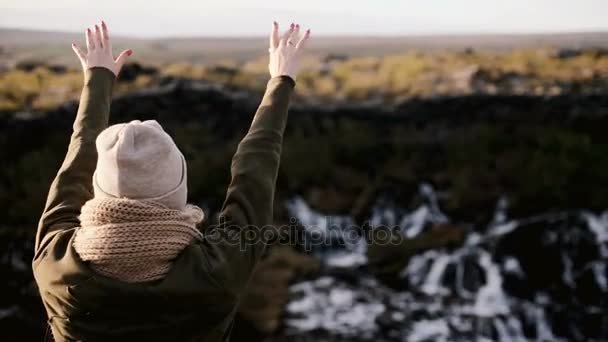 Ευτυχισμένη γυναίκα στέκεται κοντά στον καταρράκτη Barnafoss στην Ισλανδία και το άλμα. Νεαρή γυναίκα παίρνει απογειωθεί το καπέλο και σηκώνει το χέρι. - Πλάνα, βίντεο