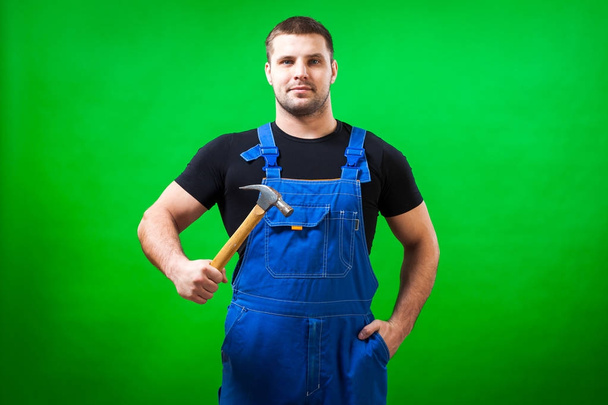 Темноволосый мужчина строитель в черной футболке и синем комбинезоне красиво позирует и держит в руке деревянный молоток на зеленом изолированном фоне
 - Фото, изображение