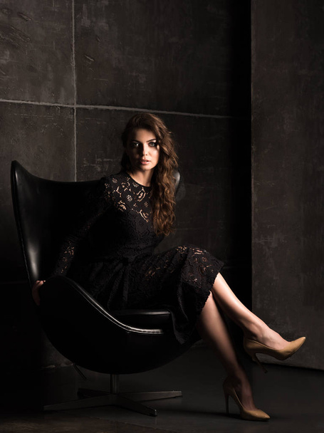 Beautiful young woman in a black lace dress sitting in a chair. Dark portrait - Photo, image