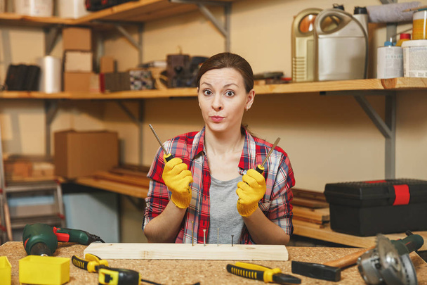 Beautiful caucasian young woman in plaid shirt, gray T-shirt, yellow gloves twisting by screwdriver screw, working in carpentry workshop at wooden table place with piece of wood, different tools. - Photo, Image