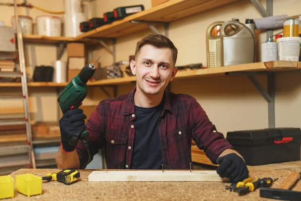 Χαμογελαστά Καυκάσιος Oμορφος νέος σε καρό πουκάμισο, μαύρο T-shirt, τοποθετήστε γάντια διάτρηση με τρυπάνι δύναμης σε κομμάτι ξύλου, εργάζεται στο ξυλουργείο στο ξύλινο τραπέζι με διαφορετικά εργαλεία. - Φωτογραφία, εικόνα