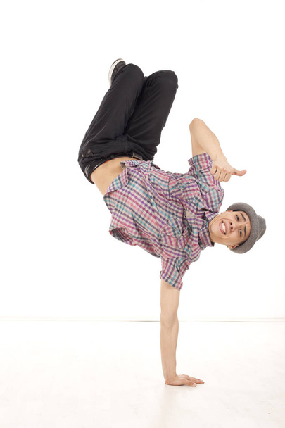 Happy young man wearing casual shirt dancing sitting on one hand, performing breakdance moves on wood floor upside down, with legs up. OK hand gesture and smile. Vertical image in studio on white background. - Photo, Image