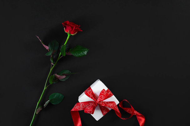 Yksi tummanpunainen ruusu punaisella nauhalla ja lahjapakkauksella mustalla backgroulla
 - Valokuva, kuva