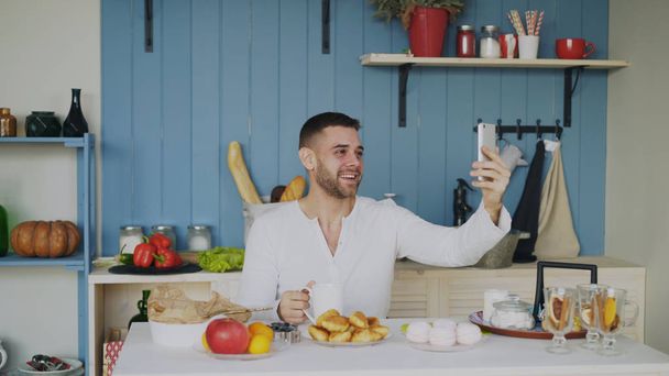 Cheerful young man using smartphone for online video chat with girlfriend while have breakfast in the morning - Фото, изображение