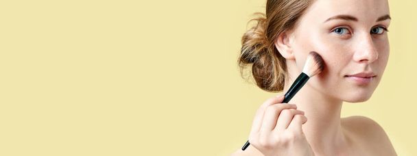 Beautiful young redhead woman with freckles contouring her cheekbones using make up brush. Beauty portrait on yellow background. - Фото, изображение