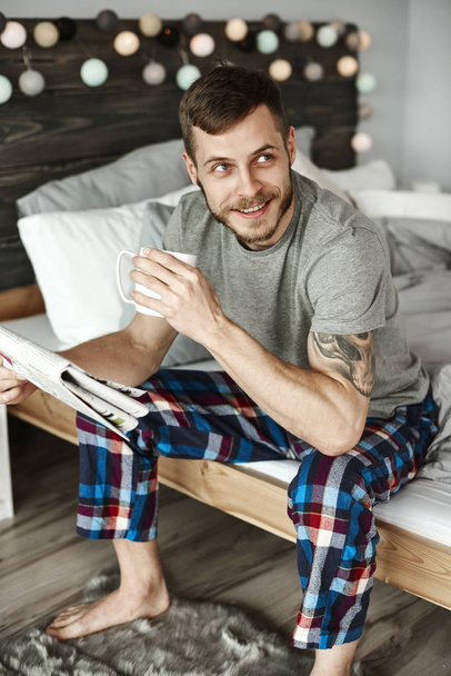 Barefoot man in pajamas with coffee sitting on bed - Photo, Image