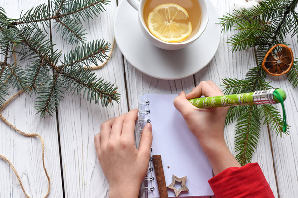 Μια γιορτινή φωτογραφία από τα χέρια των παιδιών που γράφει σε ένα βιβλίο σημείωμα, ένα cuo τσάι anf Χριστουγεννιάτικη διακόσμηση - Φωτογραφία, εικόνα