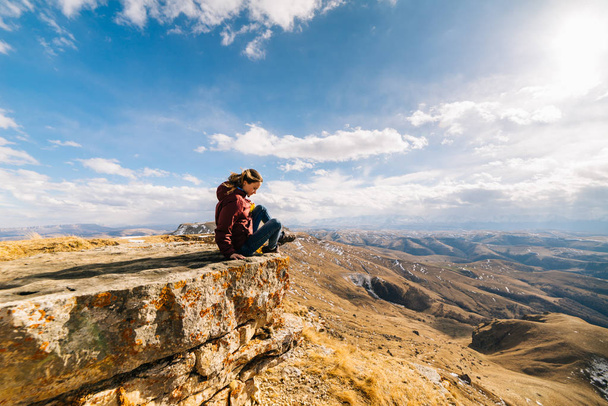 active young girl sits on the edge of the mountain, enjoys nature and the sun - Foto, afbeelding