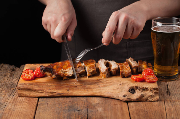 The chef cuts it with a sharp knife ready to eat pork ribs, lying on an old wooden table. A man prepares a snack to beer on a black background with copy space - Фото, изображение