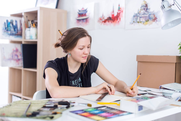 Νεαρή γυναίκα καλλιτέχνης σχέδιο σκίτσο χρησιμοποιώντας sketchbook με μολύβι στο χώρο εργασίας της στο στούντιο. Πλευρά άποψη πορτρέτο του εμπνευσμένος ζωγράφος. - Φωτογραφία, εικόνα