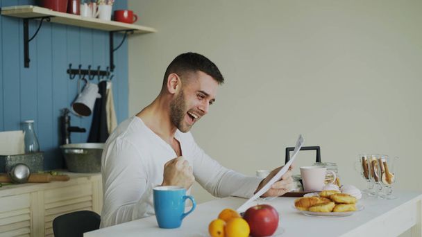 Handsome man recieve good news reading letter in the kitchen while have breakfast at home early morning - 写真・画像