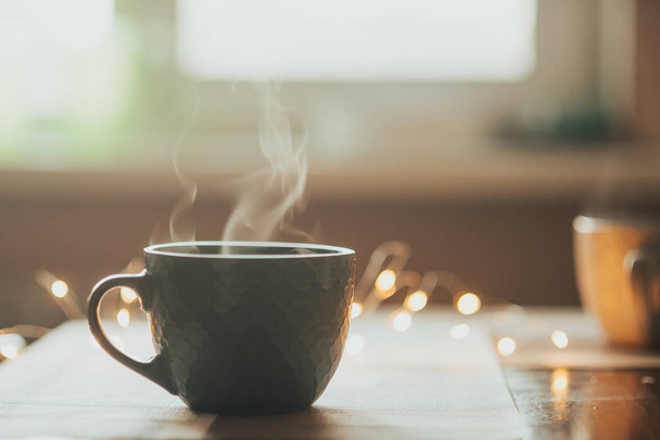Ζεστό φλιτζάνι τσάι ή καφέ. Κύπελλο με ατμό στο τραπέζι. Ζεστά τα φώτα στο παρασκήνιο - Φωτογραφία, εικόνα