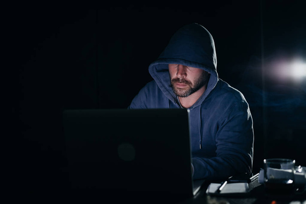 a criminal adult man hides his face under the hood, hacks the laptop - Foto, immagini