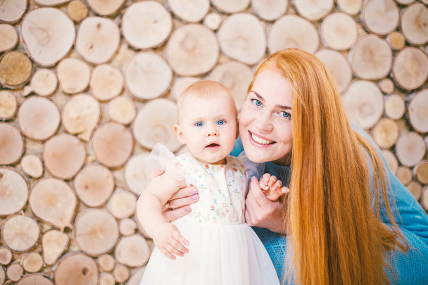 Young redhaired mom learning to walk a daughter of one-year-old at home against a background of wooden wall in the form of slices of trees. The child is dressed in a white fluffy dress.Family concept - Photo, Image