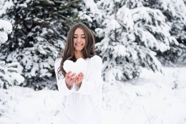 νεαρή γυναίκα στο δάσος του χειμώνα κρατά μια καρδιά από το χιόνι και να χαμογελά. Το κορίτσι δίνει την καρδιά της - Φωτογραφία, εικόνα