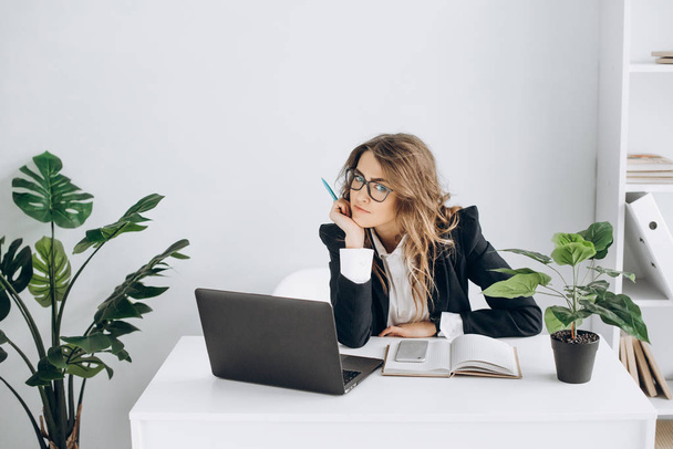 Σοβαρή επιχείρηση γυναίκα σε ένα γυαλιά και με κοστούμι, με ένα laptop κάθεται στο χώρο εργασίας της σε ένα φωτεινό γραφείο και εξετάζει τη φωτογραφική μηχανή - Φωτογραφία, εικόνα