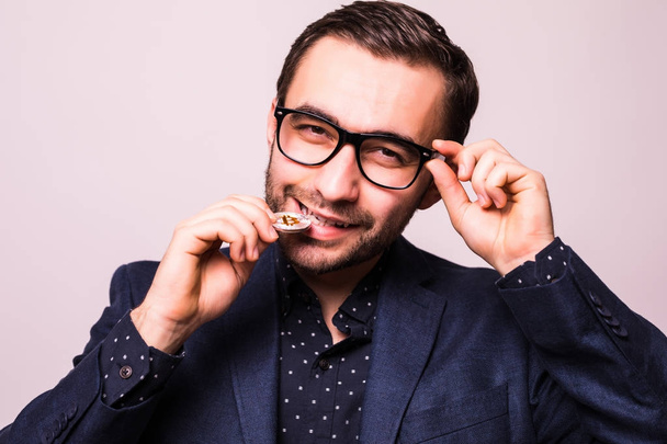 Young handsome business man in glasses bite bitcoin cion isolated on gray background - 写真・画像