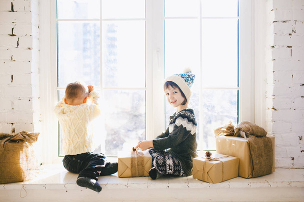 Los niños hermanos y hermanas de edad preescolar se sientan junto a la ventana en un soleado día de Navidad y juegan con cajas de regalos envueltas en papel. Están vestidos con ropa de lana caliente de punto y sombrero.
 - Foto, imagen