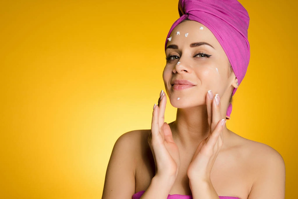 attractive young girl with a pink towel on her head applies white moisturizer on her face - Photo, image