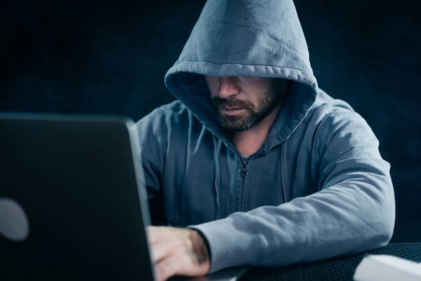 criminal bearded man hides his face under the hood, hacks the password on the laptop - 写真・画像