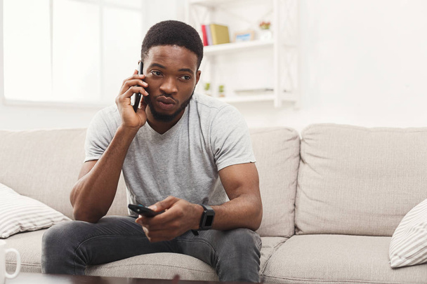 Hombre joven viendo la televisión y hablando en el móvil en la sala de estar
 - Foto, Imagen
