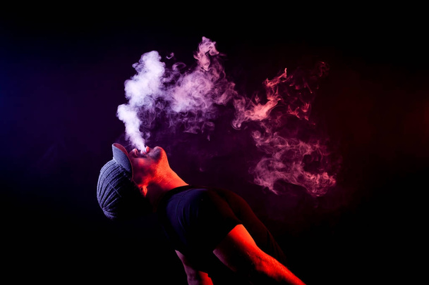 ein junger Mann atmet eine Wolke von farbigem Rauch roter Farbe auf einer schwarzen isolierten Farbe aus, die ganze Rückseite ist mit gelbem Rauch bedeckt. Schaden durch Rauchen am Beispiel eines jungen Räubers - Foto, Bild