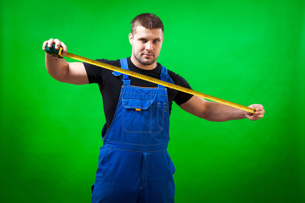 Un falegname uomo con una maglietta nera e una tuta blu usa una lunga regola del nastro giallo su uno sfondo verde isolato
 - Foto, immagini