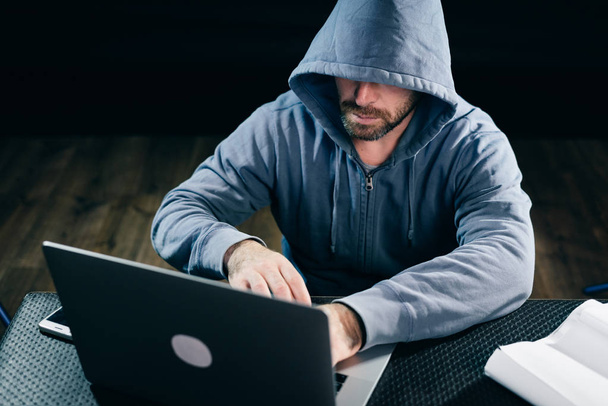 A hacker in a sweatshirt with a hood sits behind a laptop - Fotografie, Obrázek