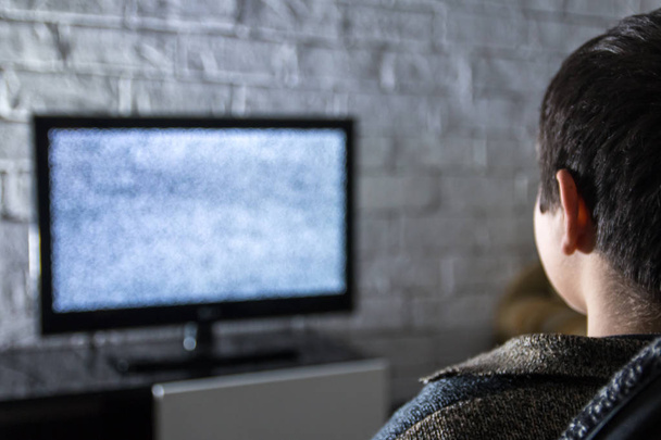 Petit garçon regardant la télévision à écran LCD dans une pièce sombre avec des murs de briques de style loft, vue de derrière
. - Photo, image