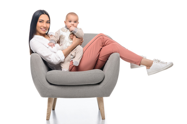 smiling young mother resting on arm chair together with little baby isolated on white - Фото, изображение