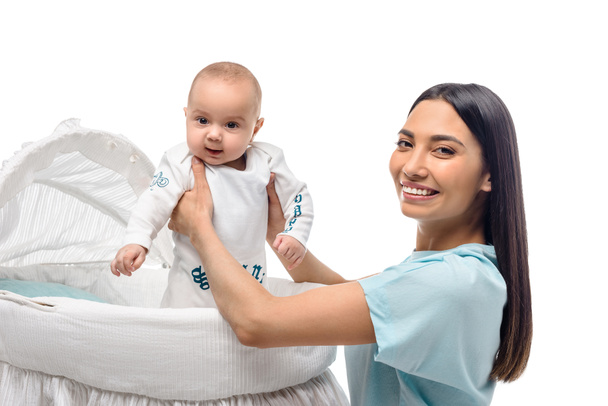 side view of smiling mother taking baby out of crib isolated on white - Photo, Image