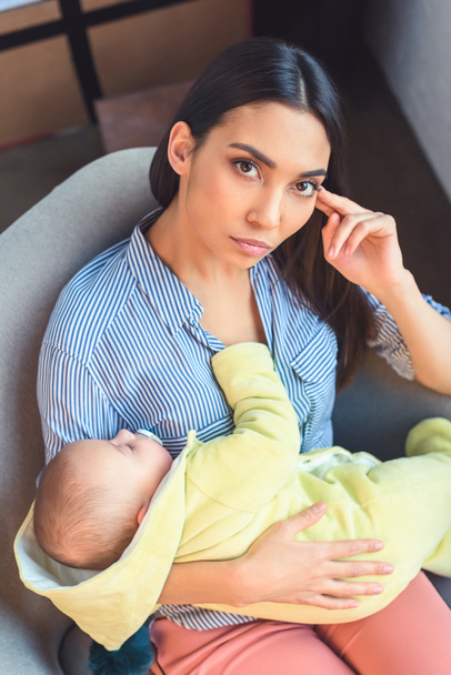 Portret van peinzende moeder met baby baby rusten op arm stoel thuis - Foto, afbeelding