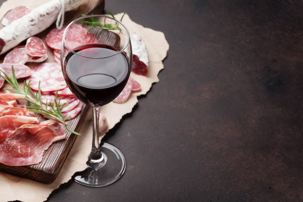 Салями, нарезанная ветчина, колбаса, прошутто, бекон и бокал красного вина. Мясное блюдо из антипаста на каменном столе. С копировальным местом
 - Фото, изображение