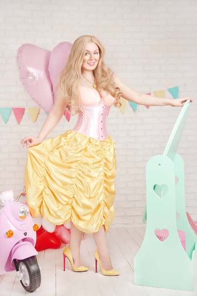 belle jolie fille blonde incurvée dans un corset rose et une jupe jaune se tient sur le miroir bleu dans la chambre blanche et amical souriant
 - Photo, image