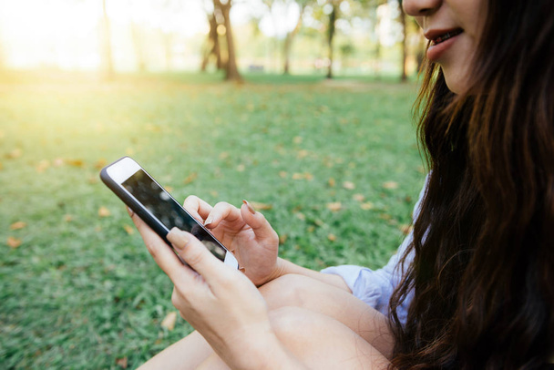 Leuke Aziatische vrouw lezen prettig SMS-bericht op mobiele telefoon zittend in park lente. Aziatische vrouw met gevoel op slimme telefoon ontspannen en smiley gezicht. Levensstijl en technologie concepten. - Foto, afbeelding