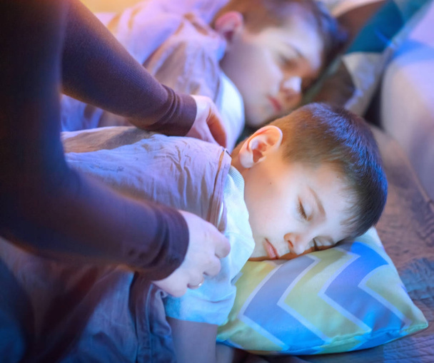 Παιδιά κοιμούνται και ονειρεύονται στο κρεβάτι, γυναικεία χέρια καλύπτοντας αγόρι με μια κουβέρτα - Φωτογραφία, εικόνα