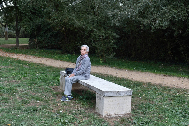 年配の女性が一人で公園のベンチに座って - 写真・画像