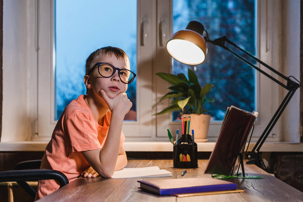 Small boy in glasses dreaming and sitting at table with lamp, books, notebooks and pens.   - Foto, imagen