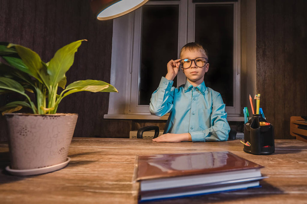 Small boy in glasses sitting at table with lamp, books, plant and pens.   - Foto, afbeelding