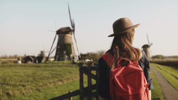 Excited tourist woman waves arms near a windmill. Traveler girl in hat with red backpack enjoys rustic mill scenery. 4K. - Imágenes, Vídeo