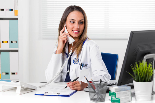 Sorrindo fêmea Doutor sentou-se na mesa com telefone para ouvido
 - Foto, Imagem