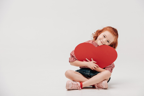 adorable little child hugging red heart symbol and smiling at camera isolated on grey  - Fotó, kép
