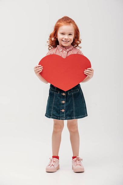 beautiful little child holding red heart symbol and smiling at camera isolated on grey  - Foto, imagen