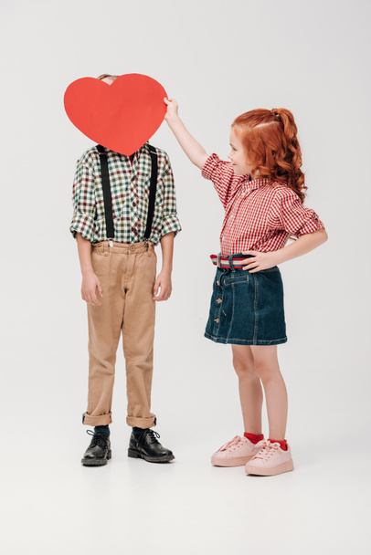 little girl holding red heart symbol near face of little boy isolated on grey  - Foto, imagen