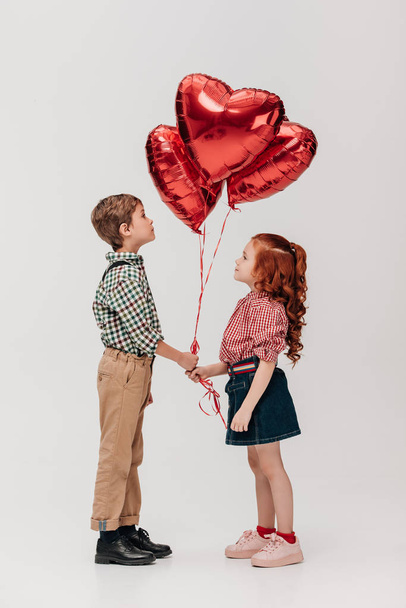 side view of beautiful little couple holding heart shaped balloons isolated on grey  - Foto, immagini