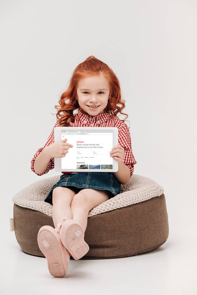 adorable little girl smiling at camera while holding digital tablet with airbnb website on screen isolated on grey - Photo, Image