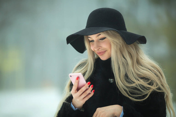 beautiful young girl in fur coat and hat walking in the park in winter weather with a phone in hands - 写真・画像