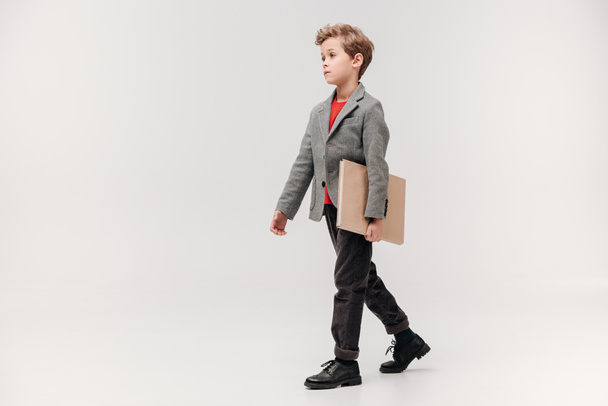 stylish little schoolboy walking with big book isolated on grey - Fotoğraf, Görsel