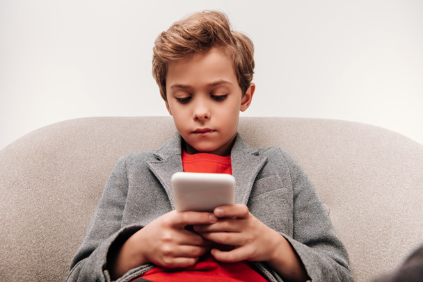 niño serio usando el teléfono inteligente mientras está sentado en sillón aislado en gris
 - Foto, imagen