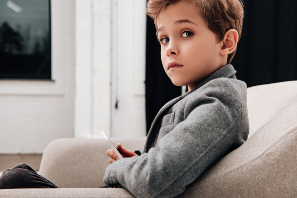 stylish little boy sitting in comfy armchair and using smartphone - Fotoğraf, Görsel